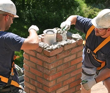 Two construction workers in hard hats and safety harnesses, from a chimney company in Long Island, repair a red brick chimney on a roof surrounded by green foliage.