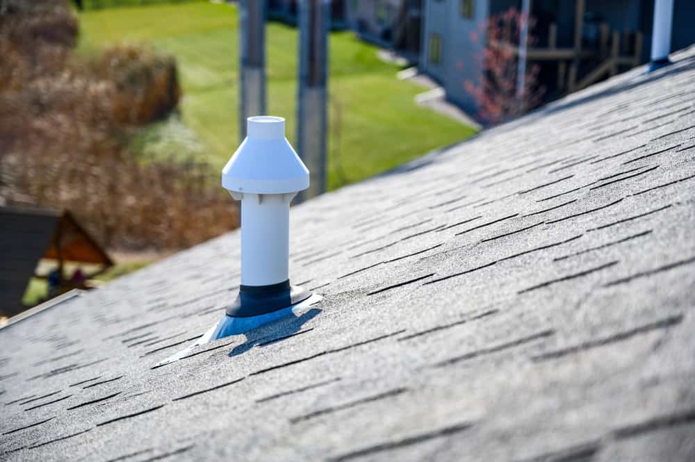 Close-up of a grey shingled roof with a white roof vent pipe in the center. Blurry green landscape and buildings are visible in the background, reflecting the expertise of a top chimney company Long Island prides itself on.