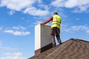A worker in a yellow safety vest stands on a roof, perhaps representing a renowned chimney company from Long Island, NY, as they expertly inspect or work on a chimney beneath a blue sky dotted with scattered white clouds.