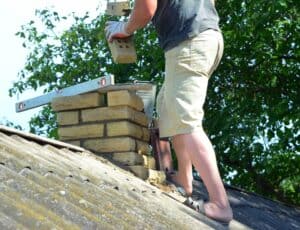 A person in shorts and sandals skillfully repairs a brick chimney on a sloped roof, holding a brick and using a metal level tool. A lush green tree basks under the clear blue sky. For quality service, consider a trusted chimney company on Long Island, NY.